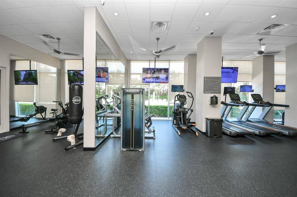 This photo shows a well-equipped fitness center with modern exercise machines, including treadmills, elliptical machines, a punching bag, and weight benches. The gym is bright and spacious, with several TVs and large windows providing natural light.