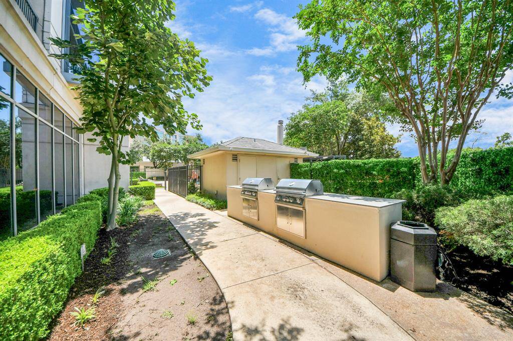 This outdoor area features a clean, paved walkway with built-in grills, surrounded by lush greenery and mature trees, perfect for entertaining and relaxation.