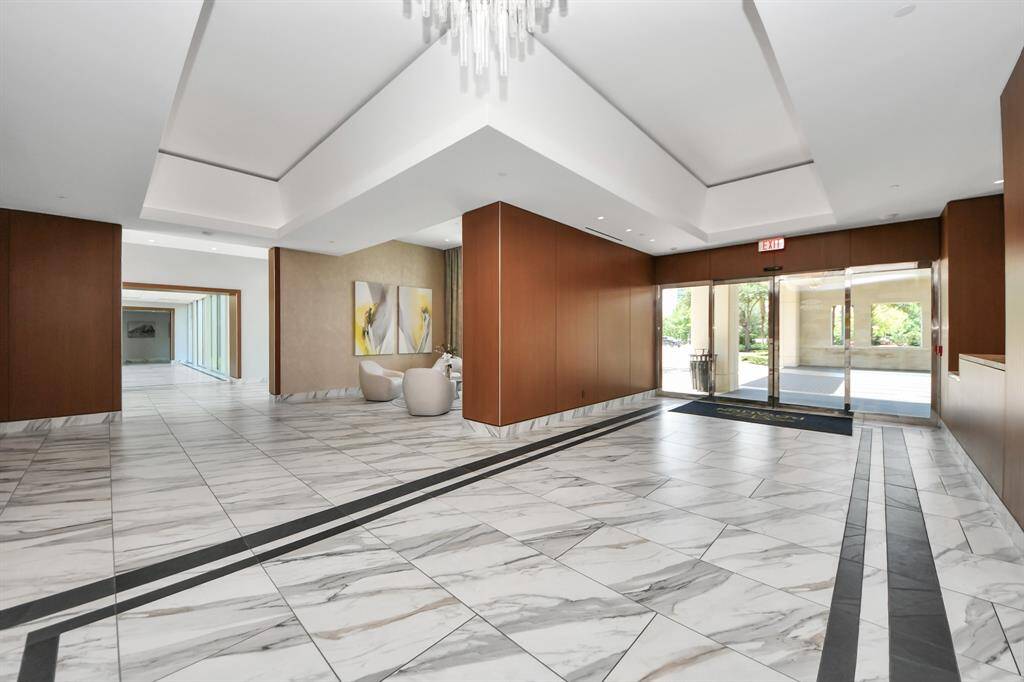 This photo shows a modern, spacious lobby with elegant marble flooring and wood-paneled walls. It features a stylish seating area, contemporary artwork, and a large chandelier. Natural light streams through glass doors, providing a welcoming atmosphere.