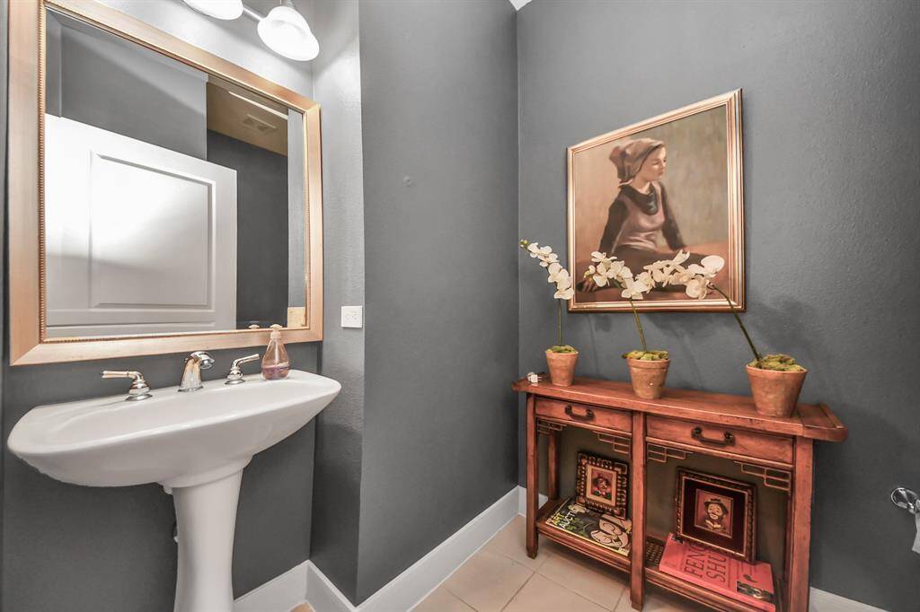 This is a stylish bathroom with a pedestal sink and a large mirror. The walls are painted in a deep gray, complemented by a wooden console table holding decorative orchids. A framed painting adds an artistic touch.