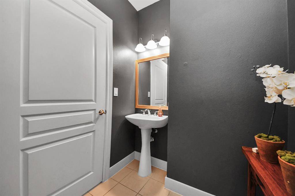 This is a modern powder room featuring dark walls, a pedestal sink with a mirror above, and elegant lighting. A decorative orchid adds a touch of nature.