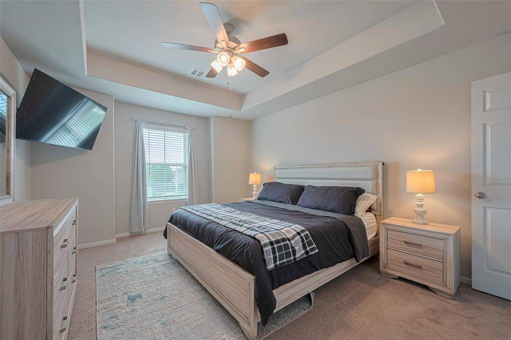 The spacious primary bedroom features soft neutral tones, plush carpeting, and a tray ceiling with a ceiling fan for added comfort.