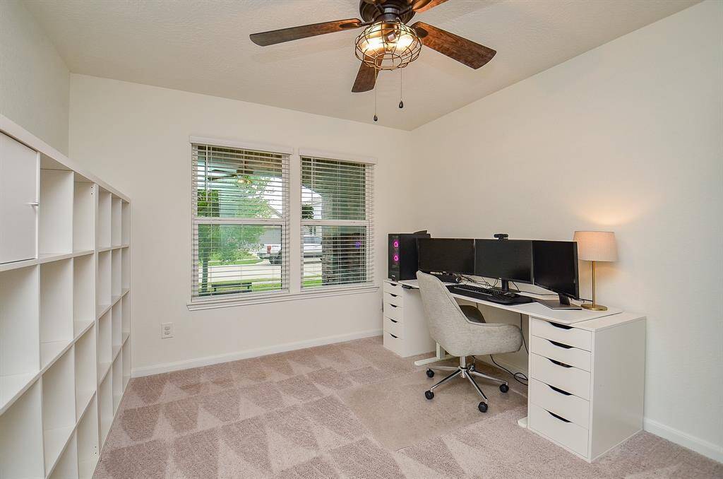 This is a bright home office with a large window, carpeted floor, and a ceiling fan. It features a modern desk setup with multiple monitors, a comfortable office chair, and ample shelving for storage. Ideal for remote work or study.