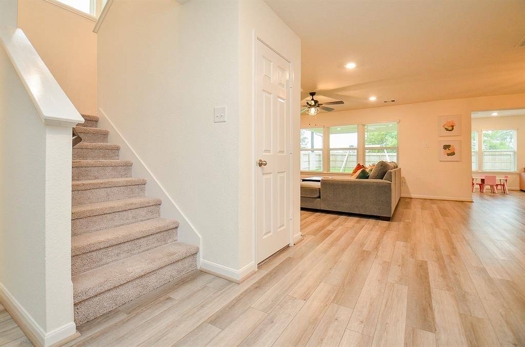This photo showcases a bright, open-concept living area with light lam. wood flooring. To the left is a carpeted staircase, leading upstairs. The living room features large windows, a ceiling fan, and a comfortable seating area. Adjacent is a dining or play area with additional windows, providing plenty of natural light.