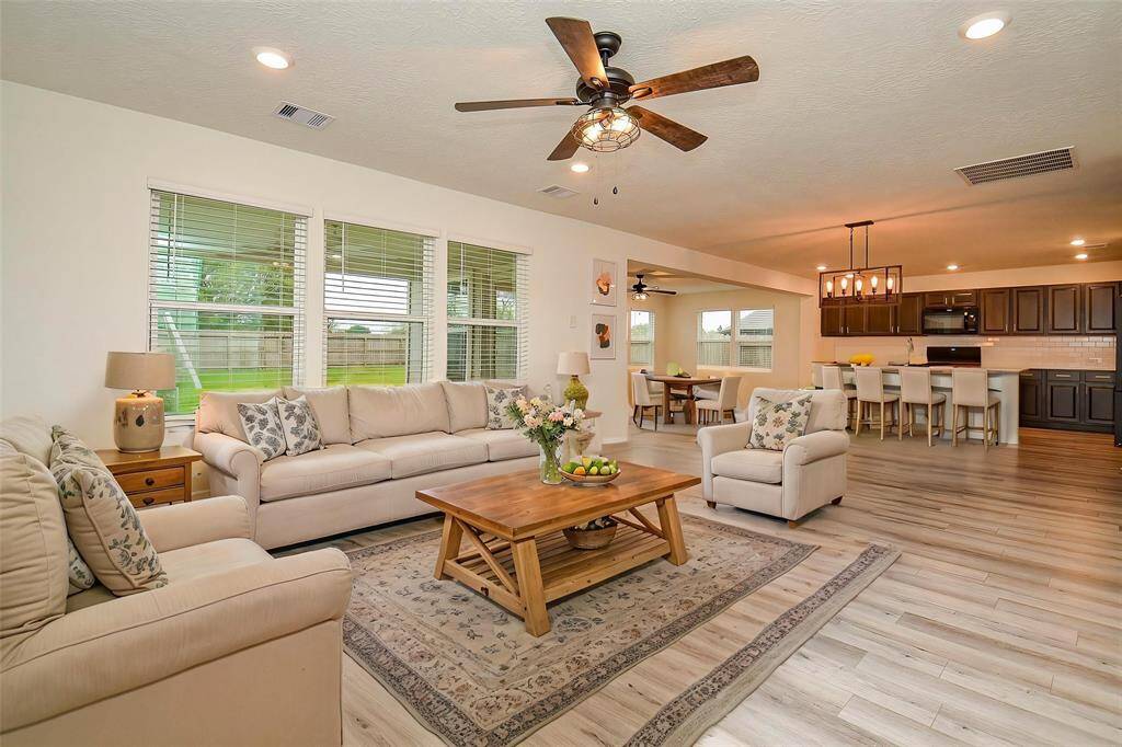 This open-concept living area features a spacious kitchen with dark wood cabinets, appliances, and a breakfast bar. The adjoining dining area has a modern light fixture and seating for six. The cozy living room is well-lit with large windows and offers a view of the backyard, creating a welcoming space for family gatherings.