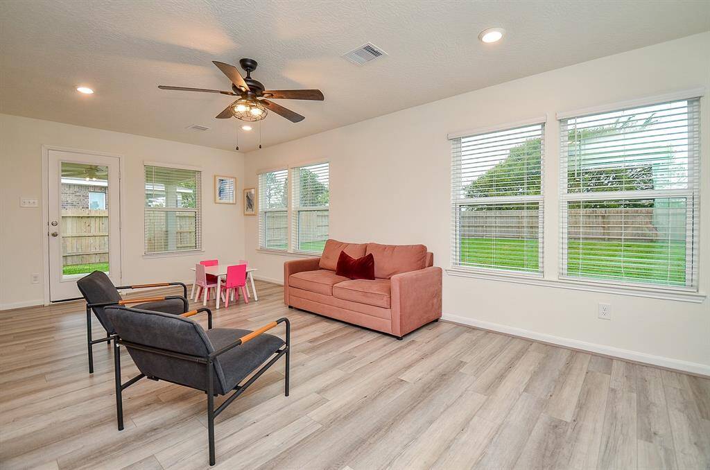 This photo showcases a spacious, open-concept kitchen with dark wood cabinets, modern appliances, and a large island with seating. The adjacent living area features natural light through multiple windows, light lam. wood flooring, and a ceiling fan.