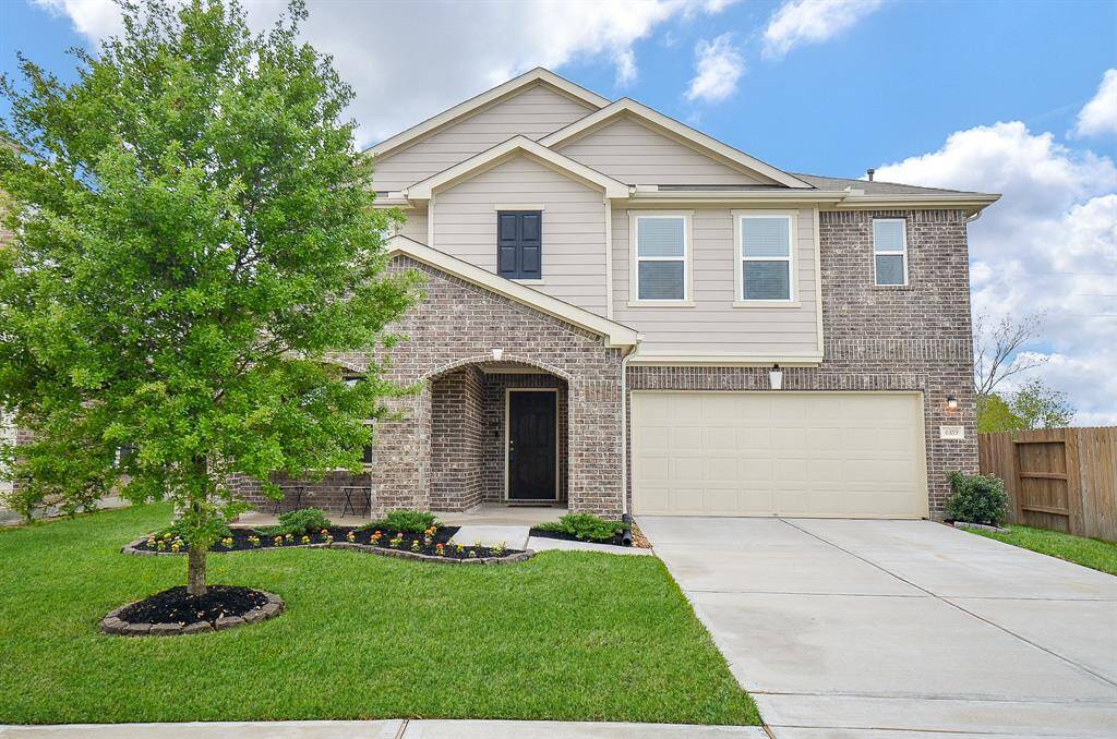 This photo shows a two-story home with a mix of brick and siding exterior. It features a neatly manicured front yard with a small tree and flower bed, plus a two-car garage. The driveway and sidewalk are well-maintained, enhancing the home's curb appeal.
