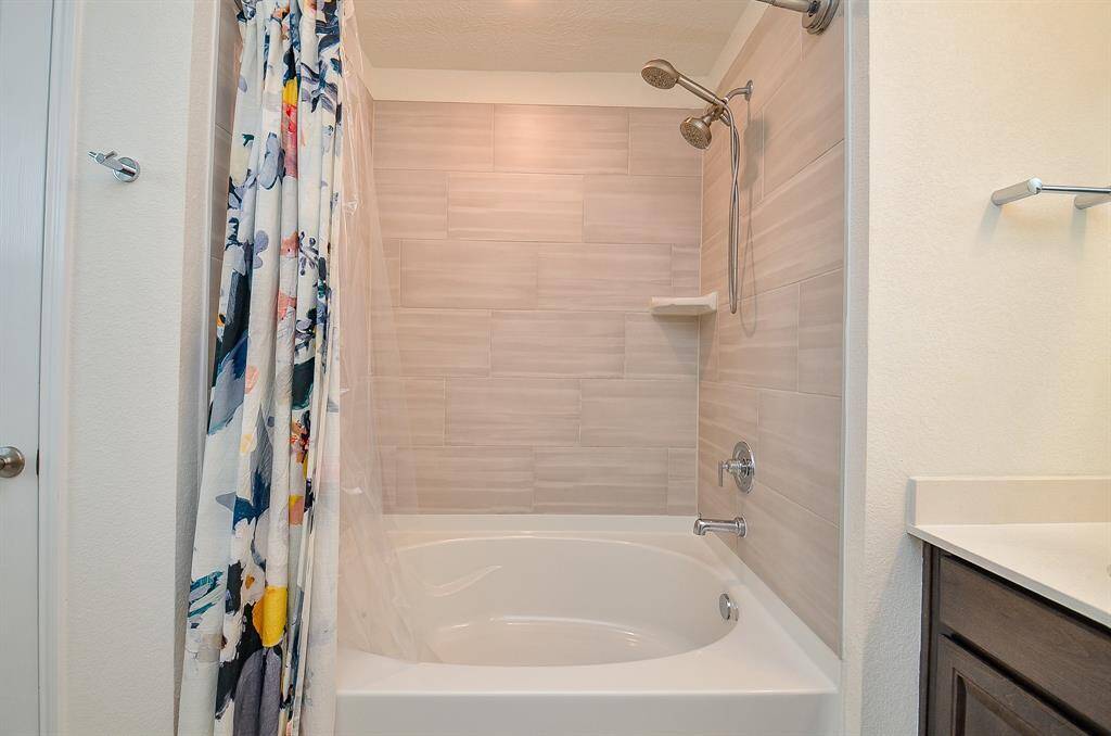 The photo shows a spacious primary suite carpeted with neutral tones. It features enough room for a large bed with a simple headboard, a ceiling fan, and modern artwork. A small nightstand with a round mirror is placed against the wall, and there’s a hallway leading to a the way to the closet and bathroom. The room feels bright and airy.