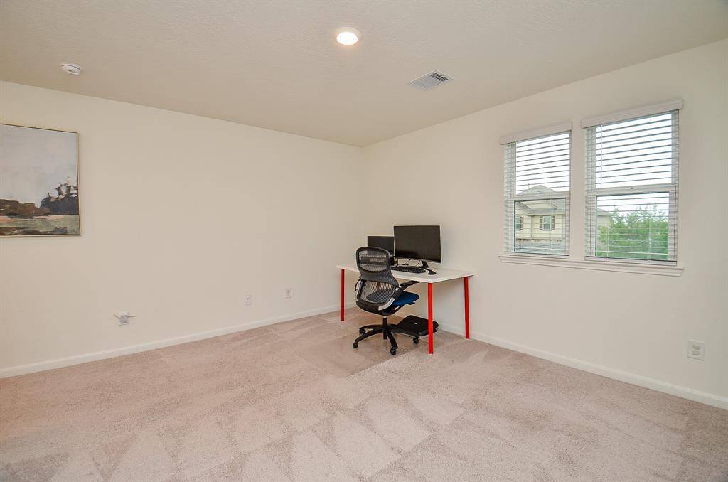 This spacious bedroom features neutral carpeting, a ceiling fan, and ample natural light through multiple windows. It includes a space for exercise equipment, creating a versatile and comfortable master bedroom area.