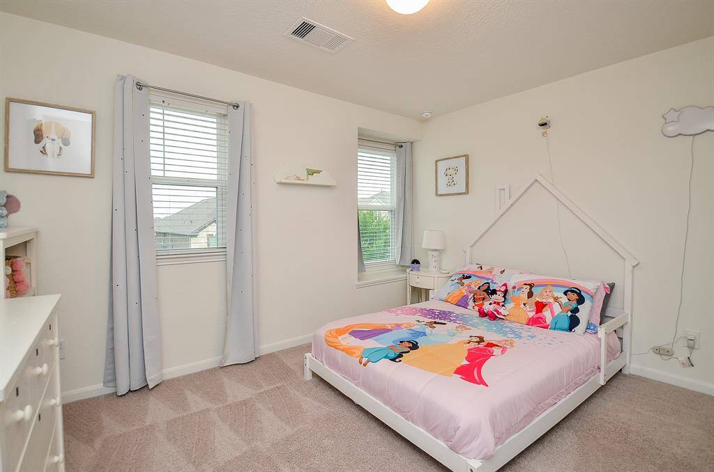 This gameroom features a simple, carpeted space with neutral walls. It includes a large window for natural light, examples of a modern desk setup with a chair, and a piece of wall art, ideal for a home office.