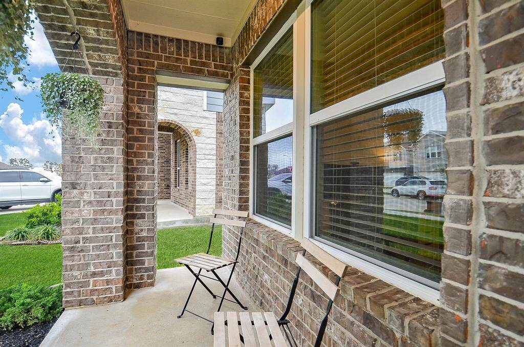 Charming brick-front porch with space for seating, featuring hanging plants and large windows, offering a cozy spot to relax while overlooking a well-maintained lawn.
