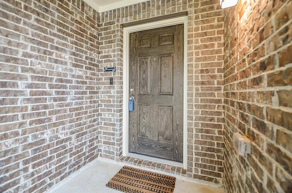 Charming entryway with a rustic, dark wood like front door framed by classic red brick walls. A welcoming touch is added by a patterned doormat, and the area is well-lit, creating a cozy first impression.
