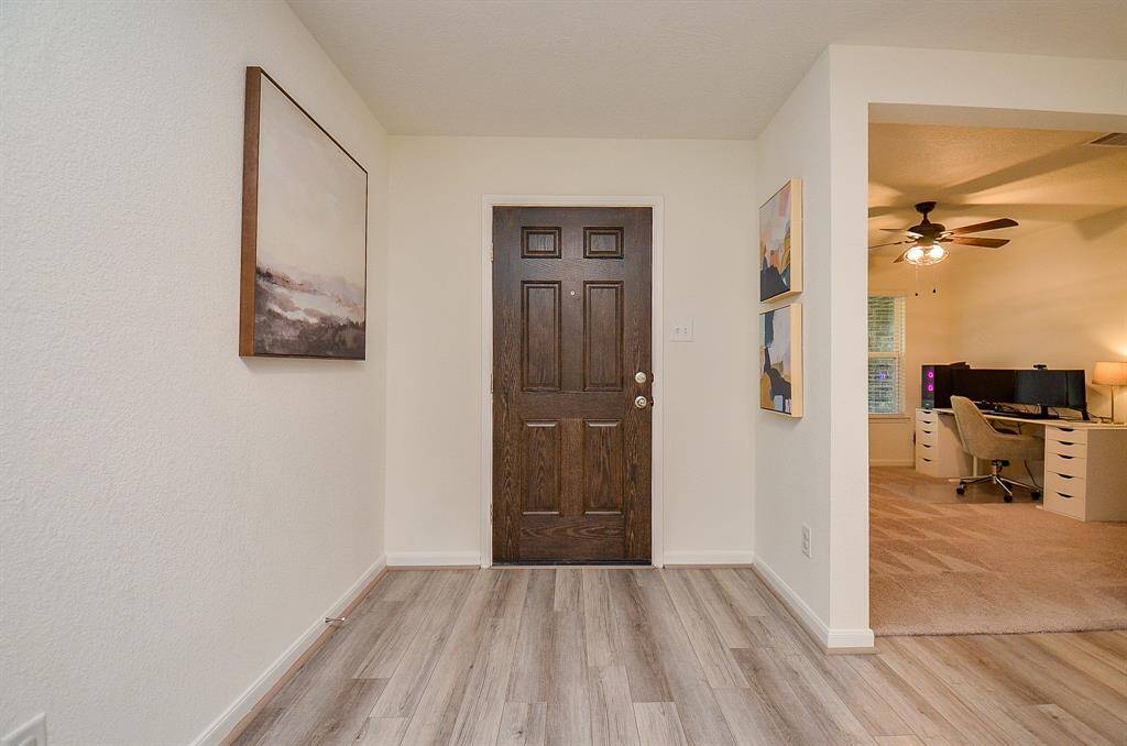 The photo shows a welcoming entryway with light laminate flooring and neutral walls. A dark wooden like door is centrally located, flanked by modern artwork. To the right, an open doorway leads to a cozy office space with carpet, featuring a ceiling fan and a workspace setup.