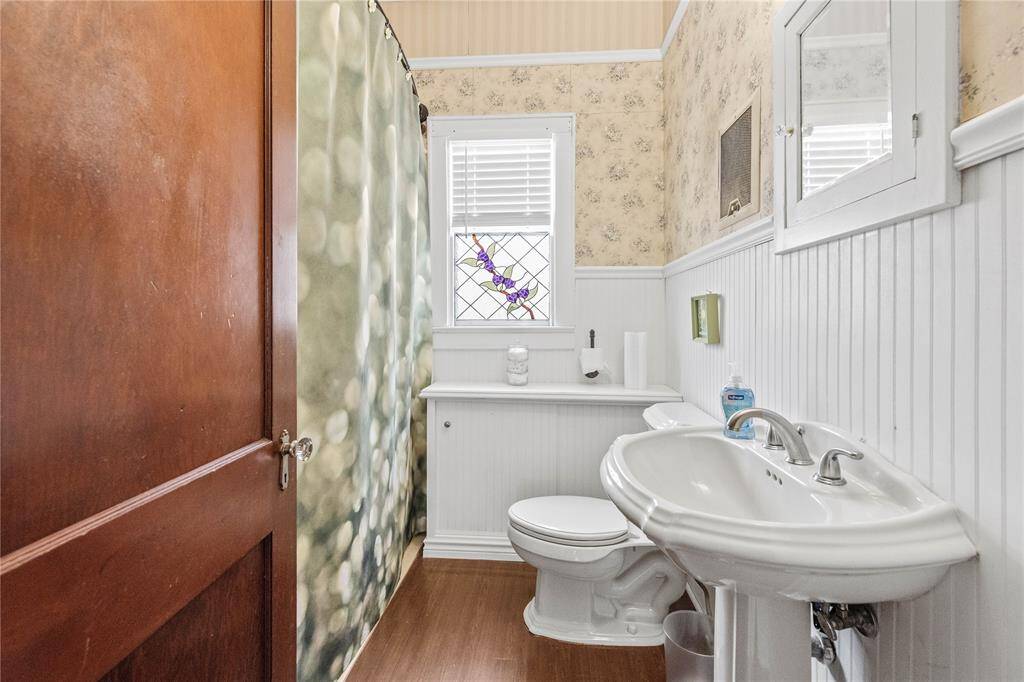 This cozy bathroom features a classic pedestal sink, a patterned shower curtain, and wainscoting with floral wallpaper. It includes a window with decorative glass, providing natural light and a touch of elegance.