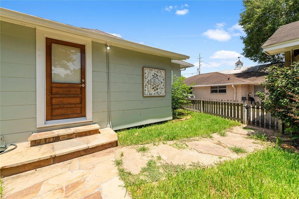 This photo shows a charming backyard space with a small grassy area and a stone path leading to a wooden door. The exterior of the house is painted a soft green, and there's a decorative wall feature. A picket fence outlines the yard, offering a cozy and inviting atmosphere.