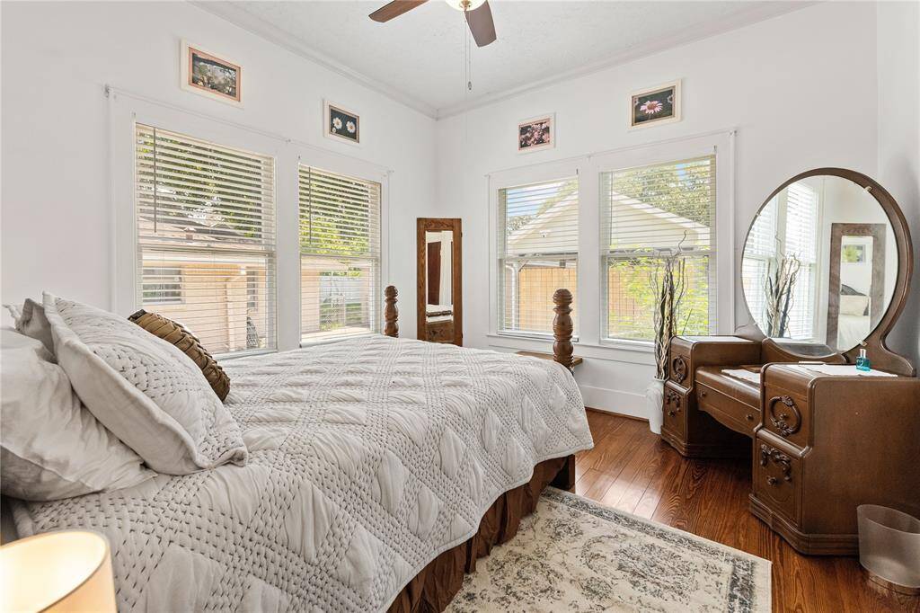 This cozy bedroom features hardwood floors, abundant natural light through multiple windows, and vintage-style furniture, including a large mirror on a wooden dresser. The neutral color palette creates a serene atmosphere.