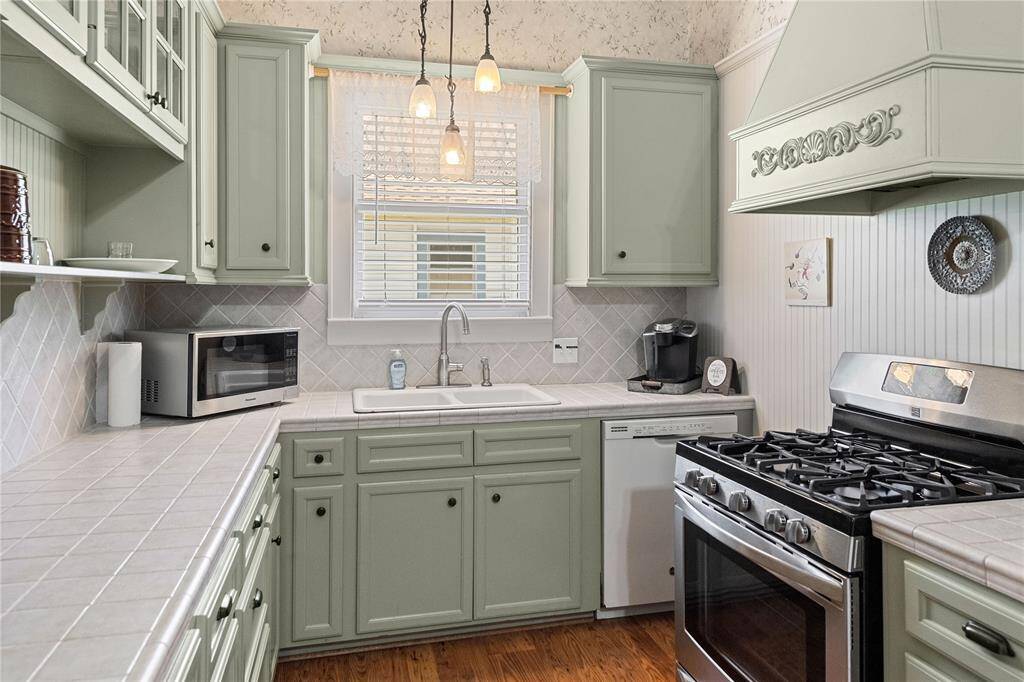 Cozy kitchen with light green cabinetry, stainless steel appliances, and a gas stove. Features a central sink under a window, pendant lighting, and ample counter space with a classic tiled backsplash.
