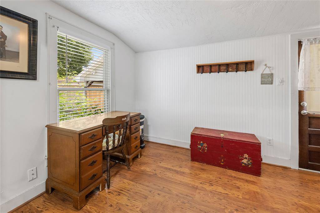 Cozy room with wooden floors, featuring a window offering natural light. It includes a vintage desk and chair, a red storage chest, and a wall-mounted coat rack. Ideal for a small office or reading nook.