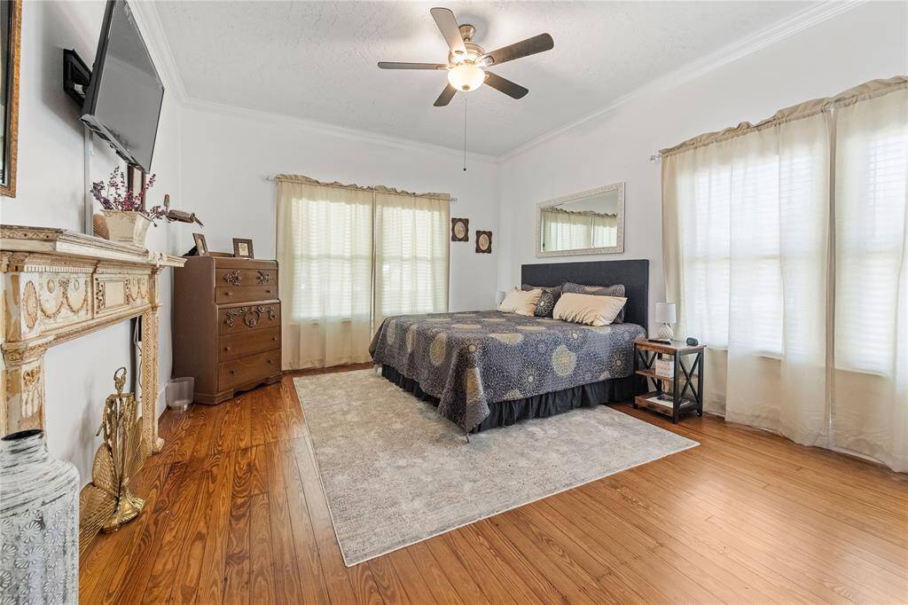 This spacious bedroom features hardwood floors, a ceiling fan, and ample natural light. It includes a decorative fireplace, elegant furnishings, and a cozy area rug, creating a warm and inviting atmosphere.