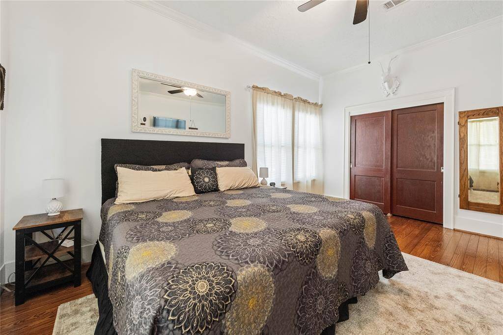 This bedroom features a king-sized bed with a decorative duvet, hardwood floors, and ample natural light from large windows. It includes a ceiling fan, a stylish mirror, and a closet with sliding wooden doors, creating a warm and inviting atmosphere.