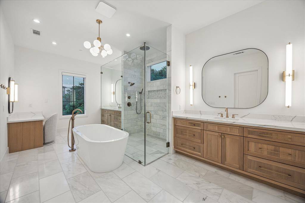 The primary bathroom is a masterpiece of refined design, featuring pristine white natural stone floors that radiate timeless elegance. Warm natural wood cabinetry adds a comforting contrast, creating a harmonious balance of texture and tone. Antique brass hardware introduces subtle vintage charm, while jewel-like fixtures bring a captivating sparkle, turning this spa-inspired retreat into a truly luxurious sanctuary.
