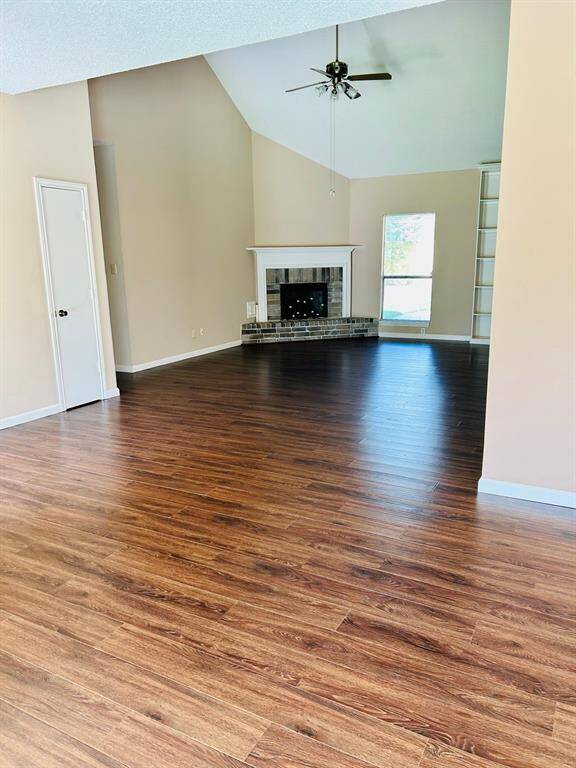 This spacious living room features high ceilings, a ceiling fan, and warm laminate wood flooring. It includes a cozy fireplace with a stone surround, built-in shelving, and a large window providing natural light.