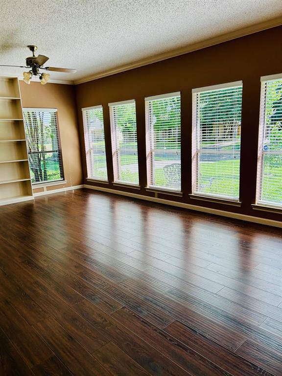 Bright, spacious room with rich laminate wood flooring, large windows offering plenty of natural light, built-in shelving, and a ceiling fan. Ideal for a living or family room with garden views.