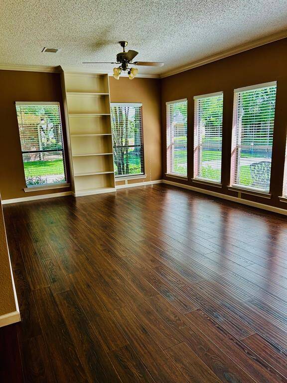This room features rich laminate wooden flooring, large windows offering plenty of natural light, built-in shelving, and a ceiling fan, creating a cozy and inviting living space.