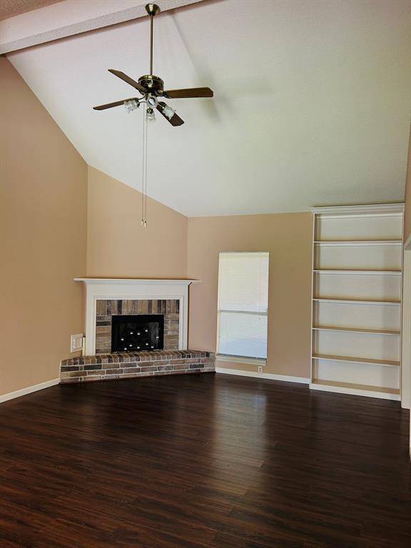 This spacious living room features 14 foot ceilings, a ceiling fan, and laminate hardwood floors. It includes a cozy brick fireplace with a white mantel, a large window for natural light, and built-in shelving for storage or display.
