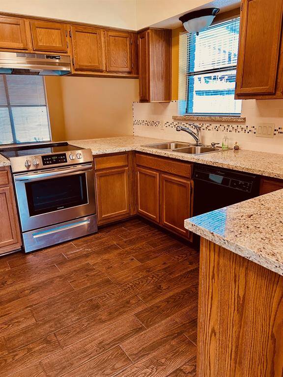 This kitchen features warm wooden cabinetry, granite countertops, and stainless steel appliances. It has a modern backsplash, dual sinks, and ample natural light from two windows. The dark wood tile flooring adds a cozy touch.