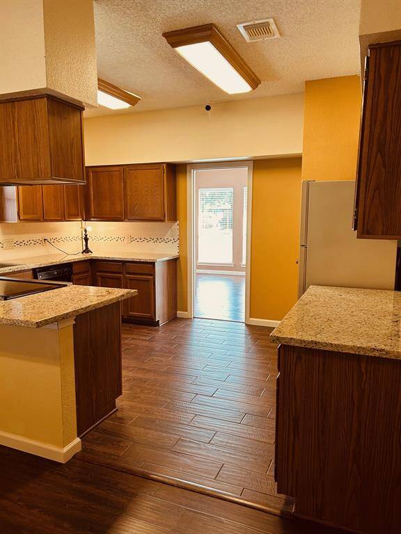 This kitchen features warm wood cabinetry, granite countertops, and tile flooring. It has ample lighting and an open doorway leading to a sunlit room, creating a bright and inviting space.
