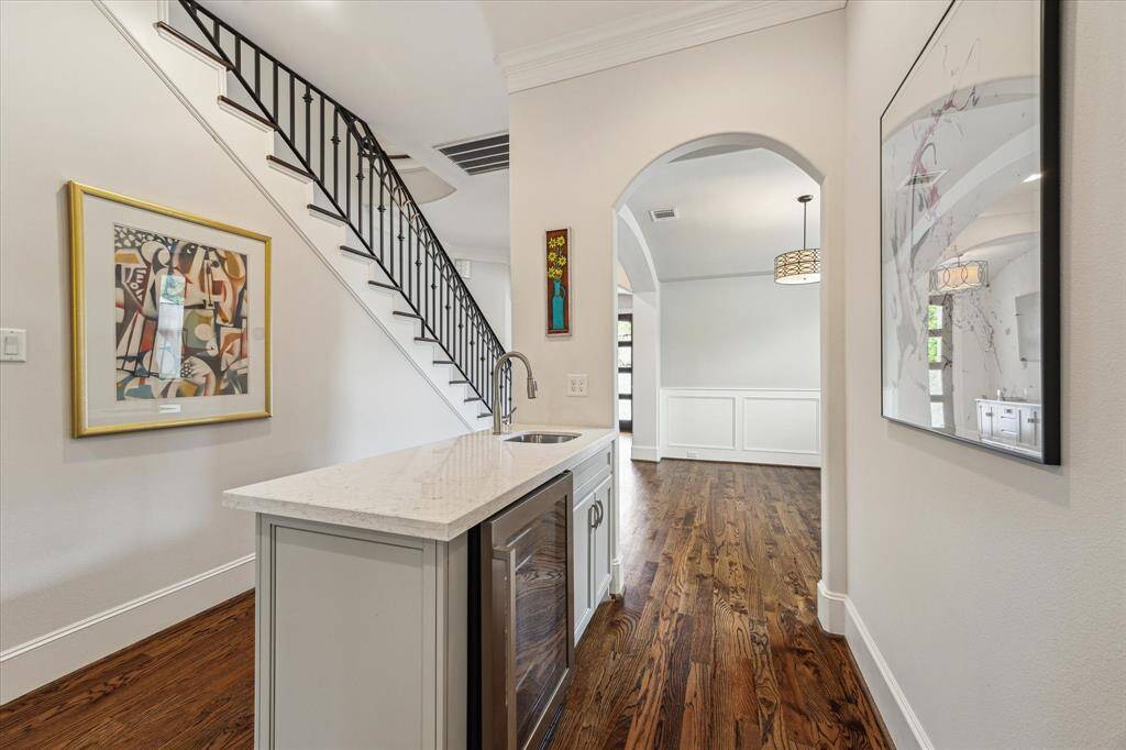 This photo showcases a stylish hallway with hardwood floors, a wet bar area with a marble countertop, sink and beverage refrigerator, elegant wall art, and a view of a staircase featuring a sleek black railing. The space is bright and modern, with an arched doorway leading to an adjoining room.