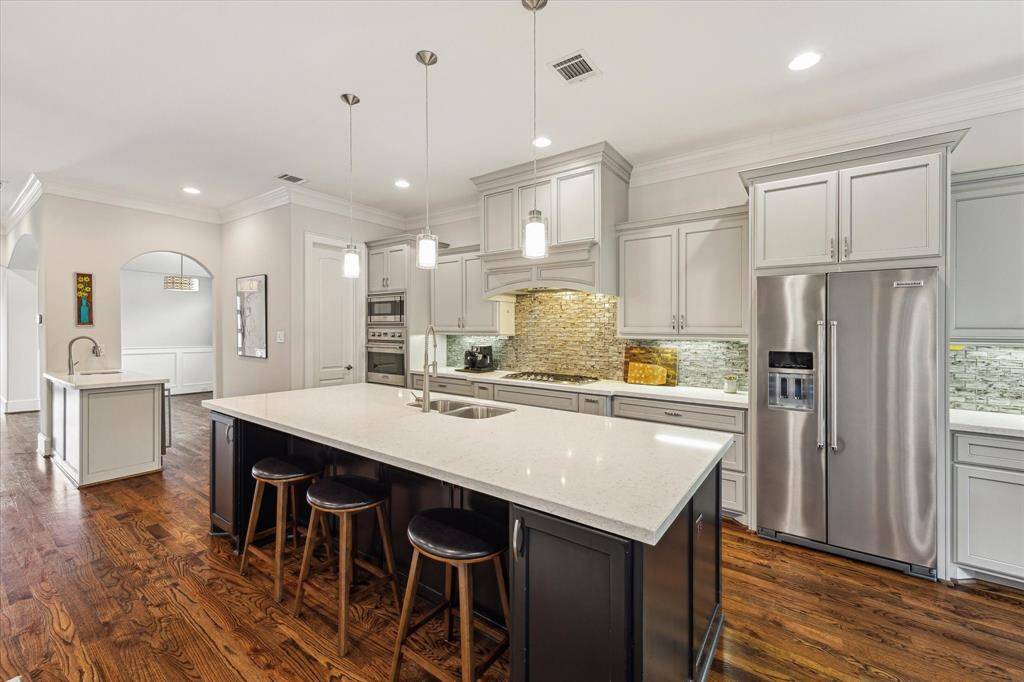 This kitchen features a spacious island with seating, modern pendant lighting, stainless steel appliances, and elegant cabinetry. The hardwood floors and stone backsplash add a touch of luxury.