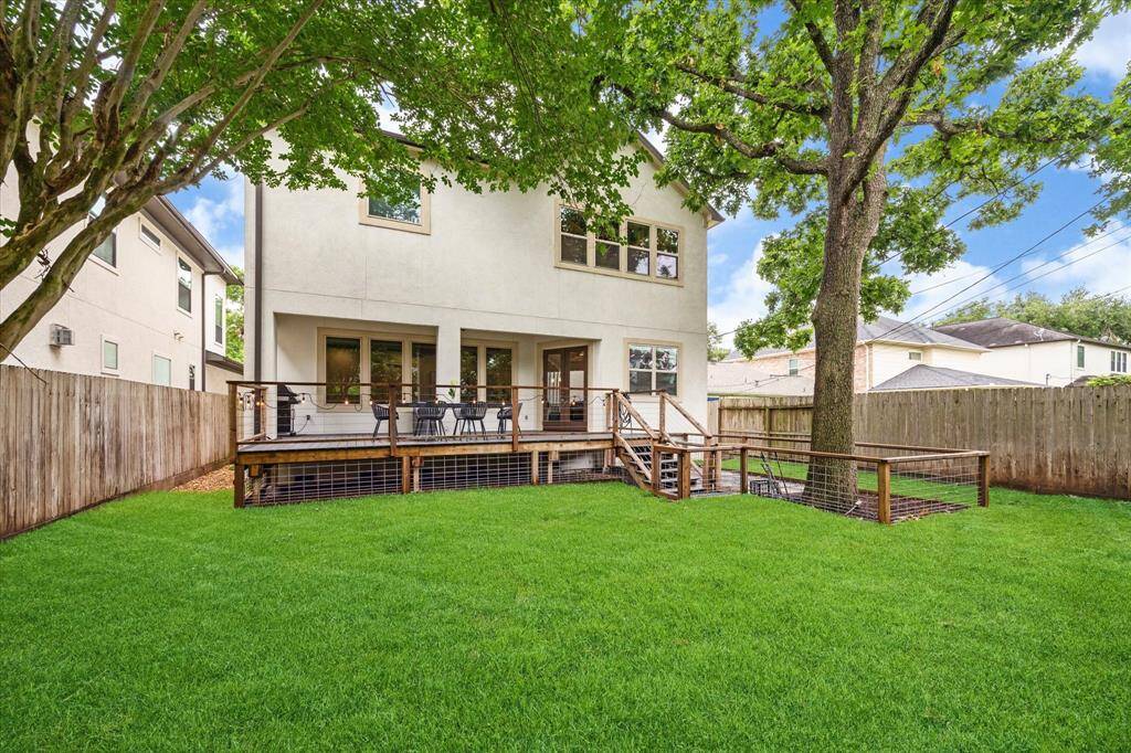 This photo shows a spacious backyard with a lush green lawn and a large tree providing shade. The back of the house features a sizable deck with outdoor seating, perfect for entertaining. The yard is enclosed by a wooden fence, offering privacy.