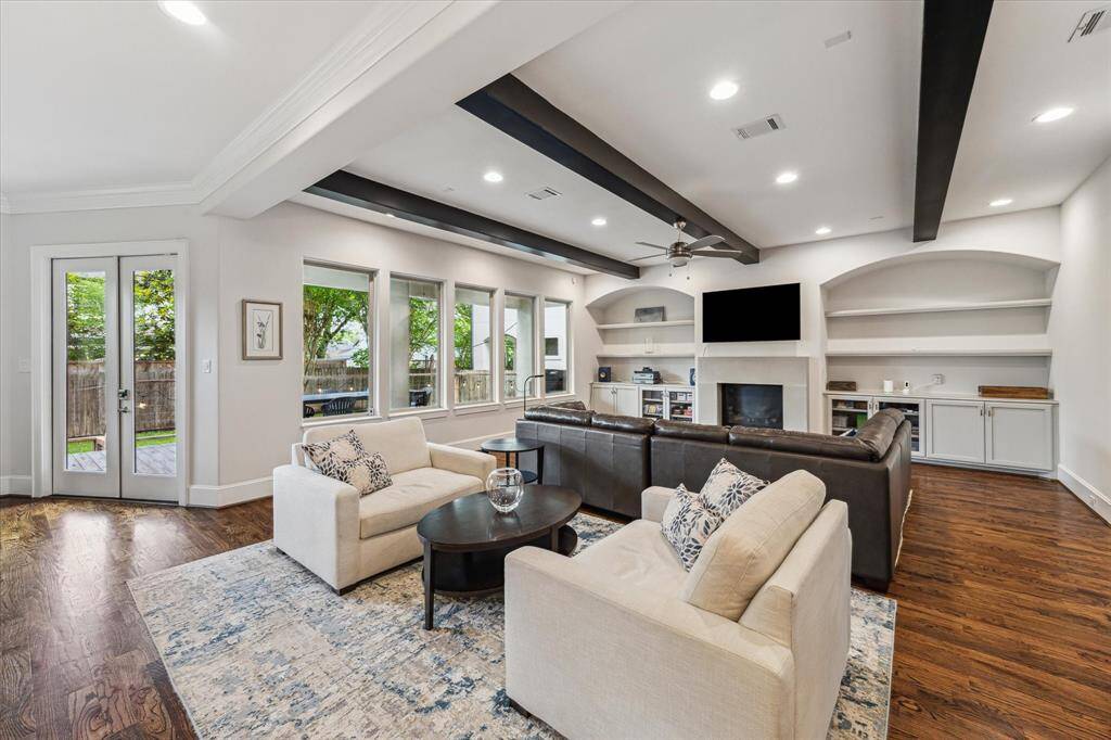 This spacious living room features elegant hardwood floors, a cozy fireplace, and built-in shelving. The room is brightened by large windows and French doors leading to a lush backyard, while dark ceiling beams add a touch of modern sophistication.