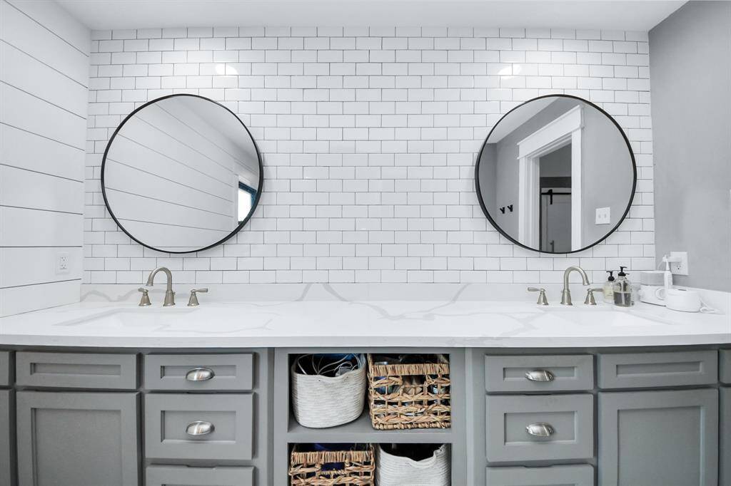 Round mirrors, a classic subway tile backsplash, soft gray cabinetry, and white quartz counters with elegant gray veining pair modern flair and timelessness in this primary bathroom.