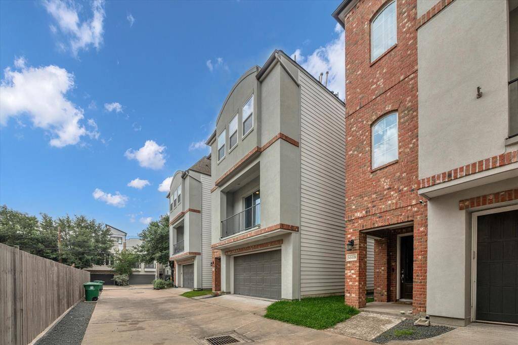 Modern single family home with brick and stucco front, featuring attached garages and balconies. The homes are situated along a paved driveway in a well-maintained area with clear skies and some greenery.
