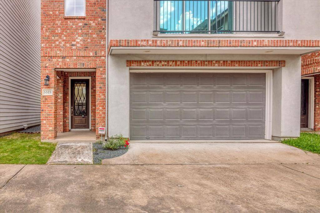 It features a two-car garage with a wide driveway and a covered porch with a decorative door. A small patch of grass and minimal landscaping enhance the curb appeal.