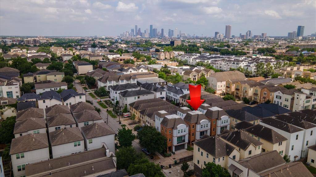 Aerial view of a residential area featuring rows of modern houses with a red arrow highlighting one property. The neighborhood is lush with greenery and set against a backdrop of a Houston skyline, indicating a convenient urban location.