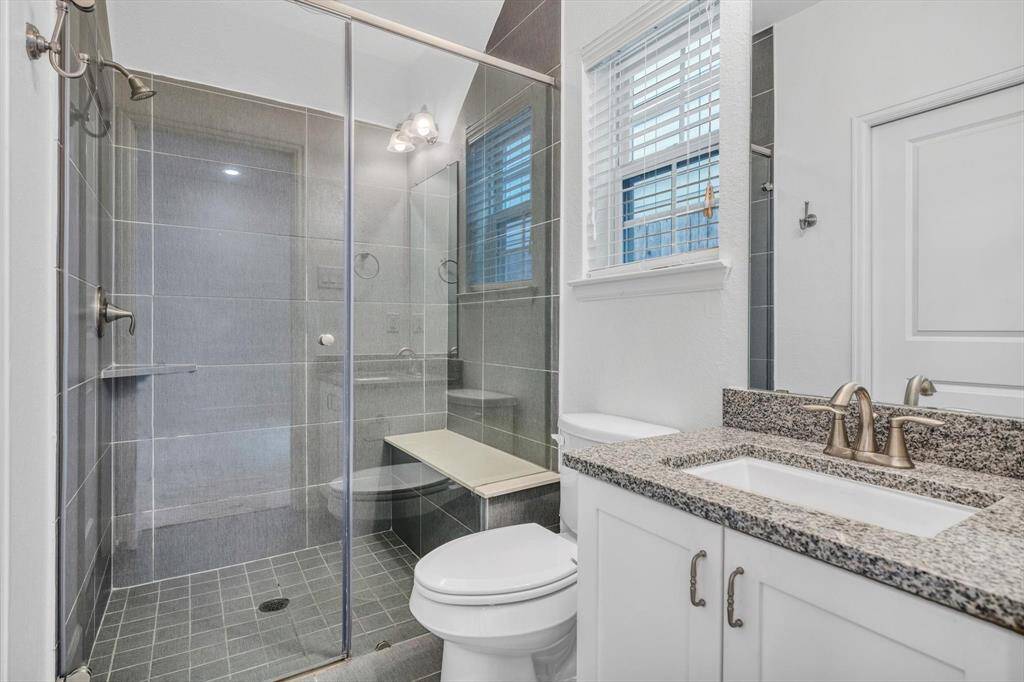 En-suite modern bathroom features a spacious glass-enclosed shower with a built-in bench, sleek gray tiles, and a granite countertop with a stylish sink. Natural light filters through a window with blinds, creating a bright and inviting space.