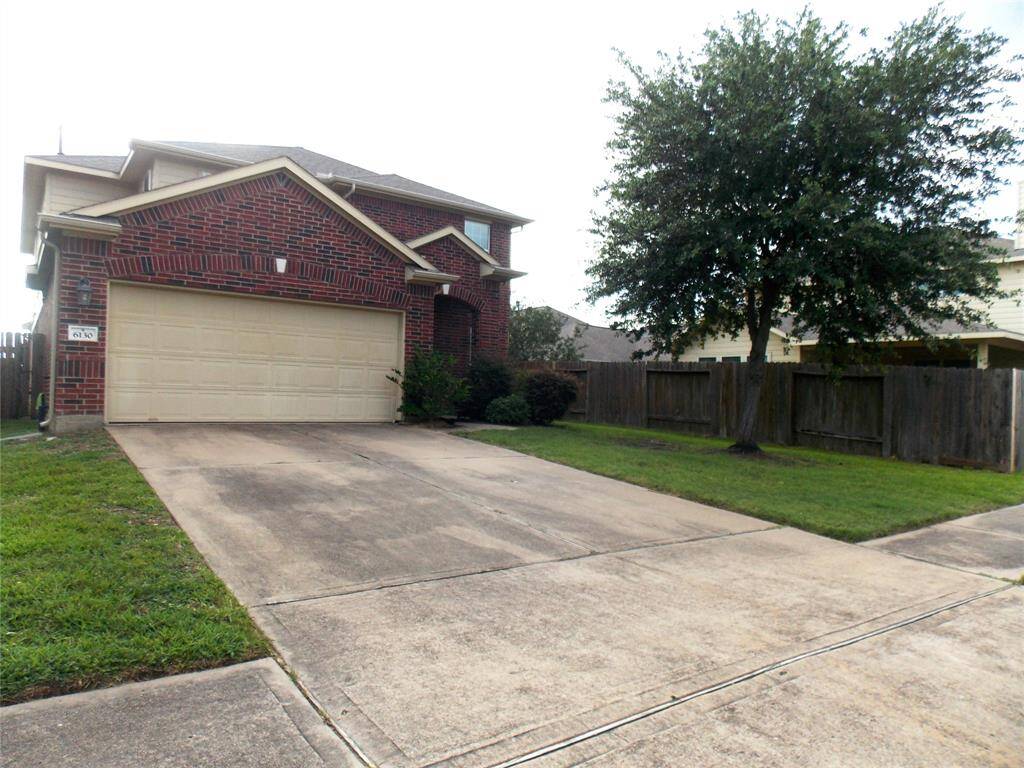 This photo shows a two-story brick house with a two-car garage. The front yard is landscaped with a tree and bushes, and there's a spacious driveway. The property is enclosed by a wooden fence, providing privacy.