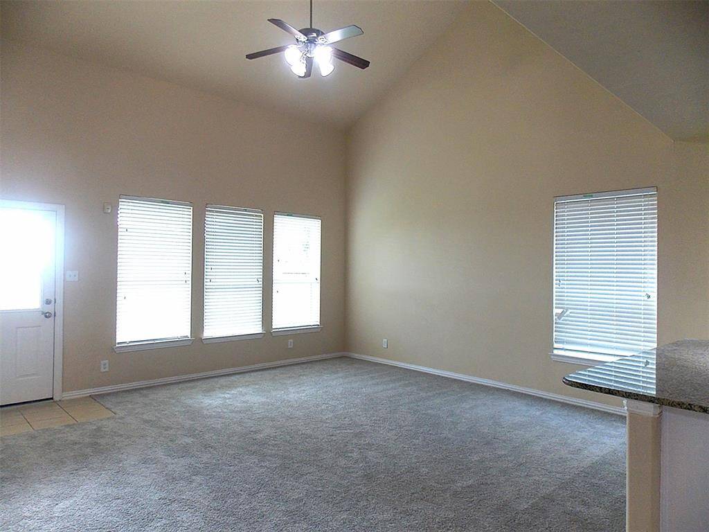 This spacious living area features high ceilings with a ceiling fan, an open layout connecting to the kitchen with a granite countertop, and a staircase leading to a loft or second level. Neutral tones enhance the bright, airy feel.