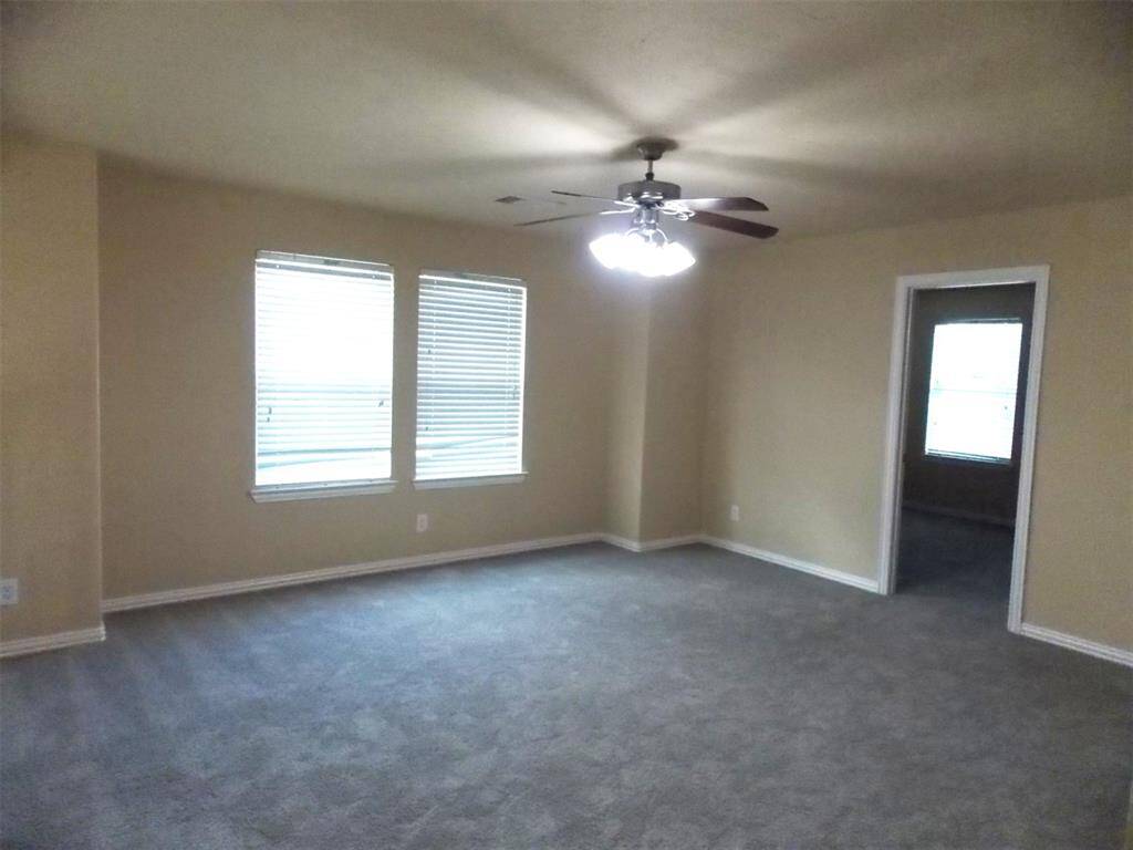 Spacious living area with high ceilings, neutral walls, and carpeted flooring. Features multiple windows with blinds and a ceiling fan. Adjacent to a kitchen counter with granite-like surfaces. Ideal for natural light and open feel.