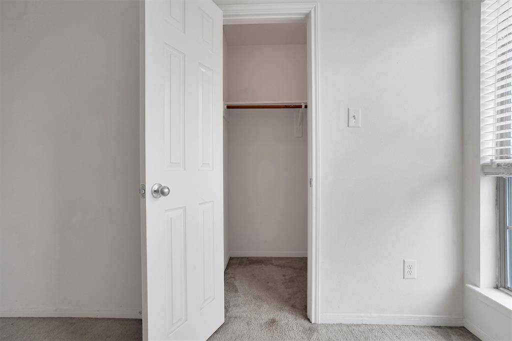 This photo shows a small, open closet with a white door in a room featuring neutral walls and carpeting. A window with blinds allows natural light to brighten the space.
