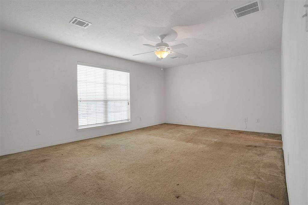 This room features neutral walls, a large window for natural light, and a ceiling fan. The carpeted floor shows some wear, but the space is well-lit and spacious, offering potential for customization.