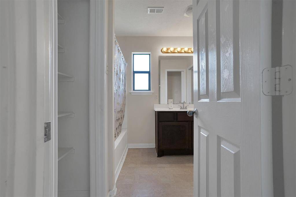 This photo shows a well-lit bathroom with a single-sink vanity, a mirror with overhead lighting, a bathtub, and shelving for storage. The neutral colors and natural light from the window create a welcoming atmosphere.