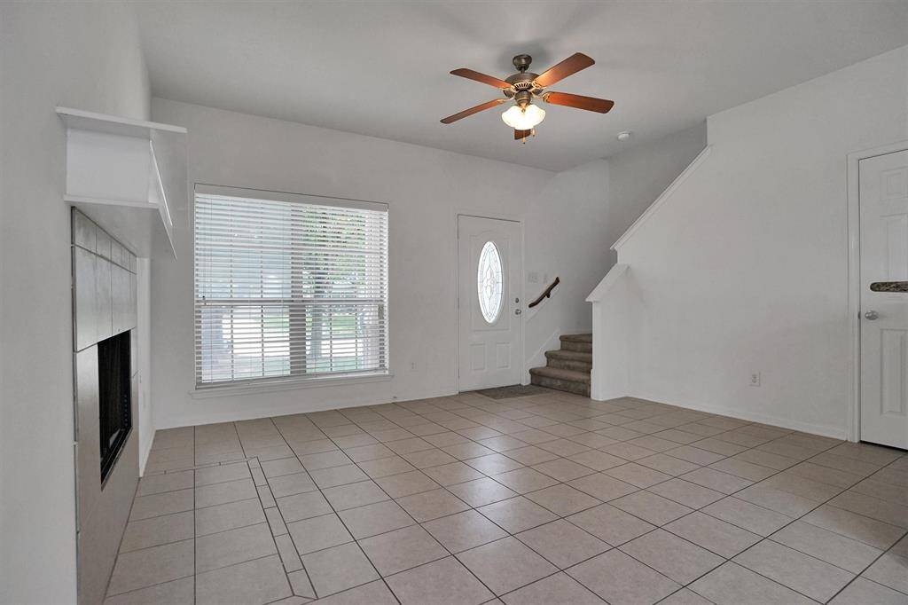 This spacious living room features tiled flooring, a ceiling fan, a cozy fireplace, and plenty of natural light from a large window. The room also includes a staircase leading to the upper level, offering a welcoming and functional layout.