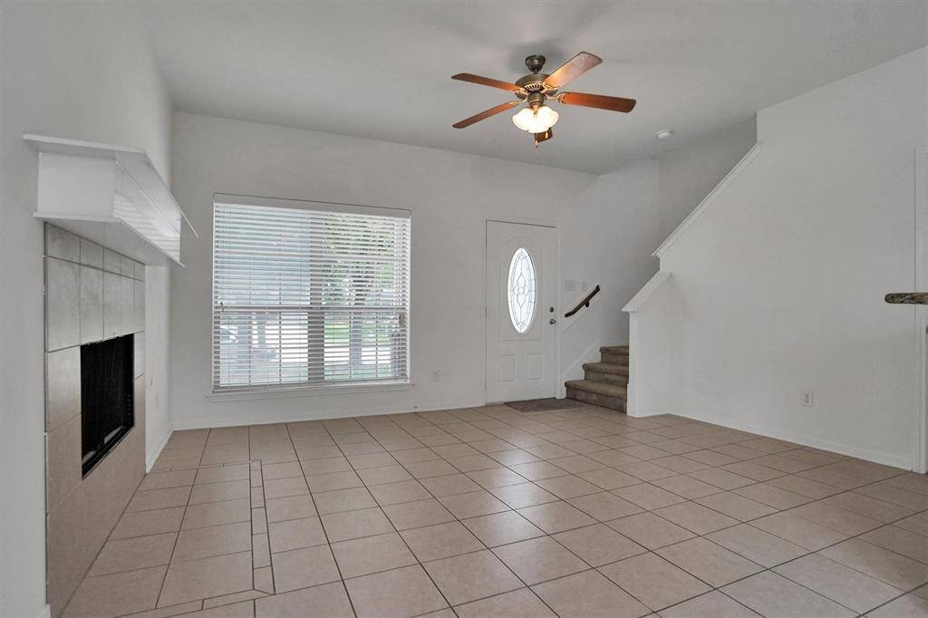 The photo shows a bright, open living space with tiled flooring, a ceiling fan, a fireplace, and a staircase. Large window allows natural light, and there's an entryway with a decorative door.