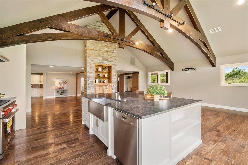 This view captures a look at the huge Island finished in designer level granite and completed with a stainless farm sink and whisper quiet dishwasher. Also, notice how the Kitchen is separated from the Grand Foyer by the floor-to-ceiling stone wall with custom built-in cabinet.