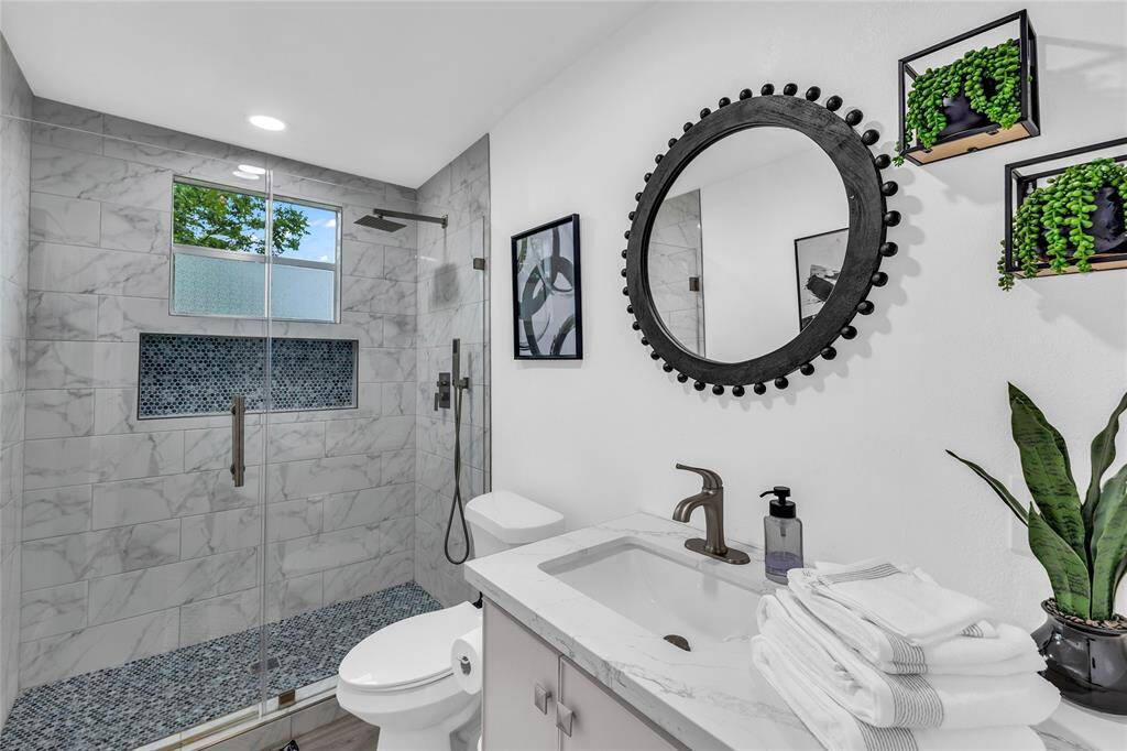 Modern bathroom featuring, a walk-in shower with glass door, white vanity, and stylish black-framed circular mirror.